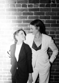 Newlyweds in black and white pant suits lean against brick wall smiling at each other