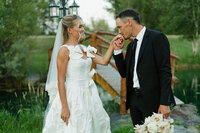 A groom kisses his bride's hand. Behind them is a wooden bridge and some greenery.