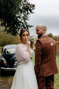 Marié observant la mariée faire le signe chut du doigt. Voiture ancienne en arrière-plan. Moment capturé par the Soulmate Duo, photographe et vidéaste de mariage.