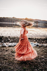 a girl twirls in the blue hour next to a the Bay water's edge.  Her hair whips around her face so that it cannot be seen.  Her twirling dress is made of an orange lace.  Her hands are on her hips.