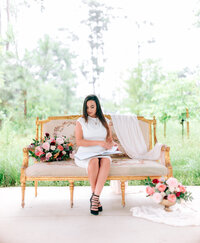 Event planner Linda Linares seated on an ornate gold sofa with pink floral arrangements, writing in a notebook at an outdoor Houston venue.