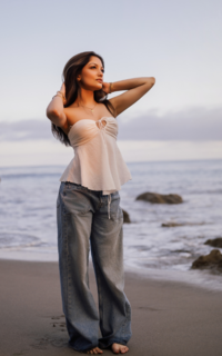 girl in white top and blue jeans at the beach holding her hands to her long dark hair and looking off to the side