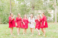 A bride in a white robe pops a bottle of champagne with her bridesmaids in a lawn wearing red robes