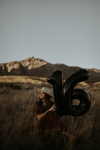 Young woman celebrating her 16th birthday in the mountains . She's holding big black number 16 balloons while the mountains are bathed in a soft glow behind her.