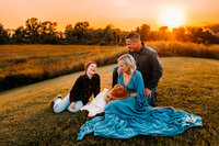 Mother father and two children sitting on the grass on top of a hill in front of a sunset