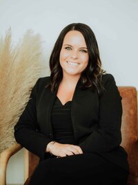 A woman with long brown hair wearing a black outfit sits smiling on a brown chair with her hands clasped, next to a decorative arrangement of light-colored pampas grass at a Maine medical aesthetics clinic.