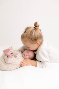 new baby wrapped in a beige wrap with older brother looking down and kissing forehead.