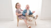 Two young siblings are seated on a small chair at Dandelion Studio in Moira, Northern Ireland. The older sister wears a floral dress and sandals, while the younger brother is dressed in a blue jacket and trousers. Both children are smiling and laughing, creating a joyful and lively moment.