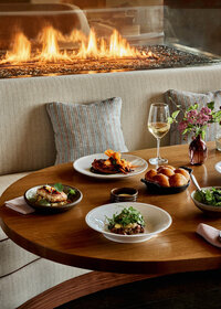 A cozy restaurant table set with various dishes, including appetizers and a main course, next to a glass of white wine. A fire burns warmly in a fireplace behind a cushioned seating area with decorative pillows.