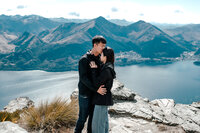 Engaged couple standing on summer cliffs in Queenstown after a helicopter proposal, overlooking the lake and mountain backdrop without snow, photographed by Zanthe Vorsatz.