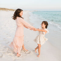 Two young sisters laugh and play while holding hands on the white sand at the beach, wearing flowy pastel dresses.