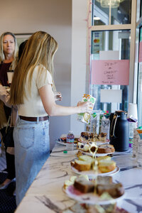 Two women stand side by side at a table filled with various cakes and pastries. The woman in the foreground is pouring juice from a carton, while the other holds a cup and looks at the table. A sign is visible in the background. There's a large window to the right, offering a view of Seattle's skyline.