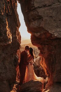 A couple shares a quiet moment nestled between two large rocks, bathed in the golden light of sunset. The scene is set on Mount Lemmon in Tucson, Arizona, with a breathtaking view of the surrounding landscape.