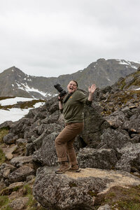 Sydney Breann, an elopement photographer, smiles and waves while holding her camera on rocky mountain terrain surrounded by snow patches in Alaska.