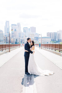 Bride and groom standing on the Stone Arch Bridge in Minneapolis posing for a photo, hugging while he kisses her head - captured by Minnesota wedding photographer. 