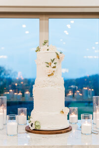 A 3 tiered wedding cake decorated in white frosting and florals, surrounded by candles at twilight