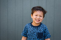 portrait of a preschool boy standing in front of a blue wall at a Bay Area school looking at the camera and smiling