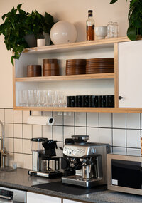 Minimalist office kitchen with espresso machine, glassware, brown dishes, and potted greenery on open shelving.