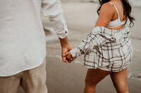  Close-up of a couple holding hands during a maternity photoshoot, with the woman wearing a plaid shirt and shorts, walking barefoot on the beach in Southern California

