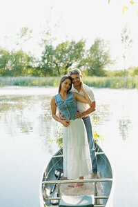 Looking and smiling at the camera, a man is holding his fiance by her waist and she is holding his face with one hand. They are standing up in a rowboat that is on a lake.
