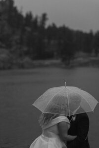 Groom kisses bride's forehead while protecting her from the rain under a clear umbrella