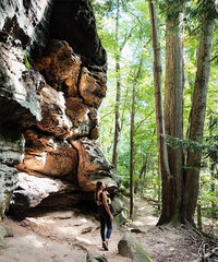 A woman explores the forest of Hocking Hills, with a unique rock formation rising among the trees nearby.