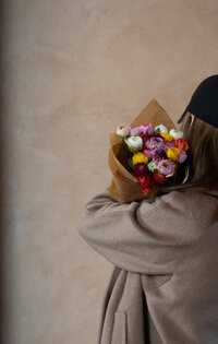 A woman wearing a brown coat and baseball hat holding a bouquet of bright flowers 