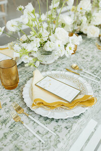 Elegant spring wedding place setting with yellow napkin, modern menu design, white florals, and amber glassware, styled by Eventive Designs in Austin.