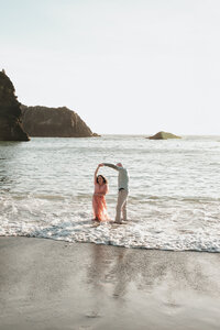 Romantic couple dancing in shallow ocean water at Secret Beach, a secluded and iconic Southern Oregon coastal cove, surrounded by rugged rocky cliffs and glowing in golden hour light, captured by a Pacific Northwest wedding photographer specializing in intimate and adventurous wedding sessions. 