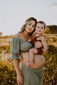 A pregnant mom holding her toddler in her arms while the boy looks at the camera in a field in fort worth, texas.