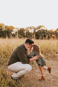 dad squatting down to kiss his daughter in a field at sunset