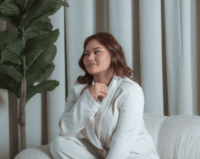 Stylish woman with shoulder-length hair wearing a white outfit, sitting comfortably on a textured couch with neutral curtains and a tall green plant beside her.