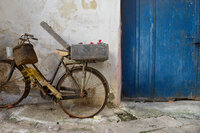 An old bicycle leans against a textured stucco wall, with a wicker basket, sprigs of wheat, and a box of red flowers. A bright blue weathered door anchors the scene, evoking rustic charm and everyday beauty in a quiet Italian village.