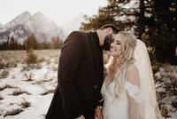 grand teton wedding in the winter with groom leaning to whisper something in his brides ear and the bride who is in a lace gown smiles with the mountains in the distance captured by jackson hole photographers