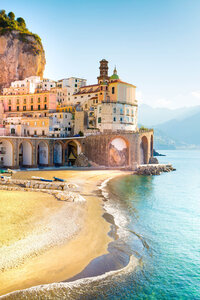 A picturesque coastal village with colorful buildings perched on a rocky cliff overlooking a sandy beach and clear blue sea. The sun illuminates the scene, casting a warm glow over the landscape.