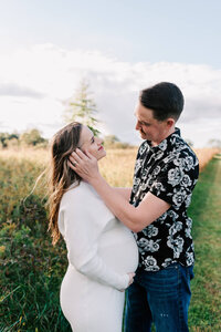 a husband in a black floral shirt is sweetly pulling the hair out of his expecting wife's face, during their maternity session in Northern VA