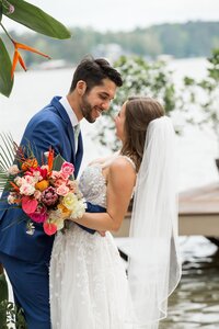Man in navy suit leaning in to women in white dress with veil, as she is holding a bouquet of pink tropical flowers. 