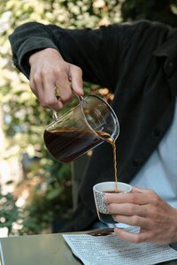 Small circular photo on a green background showing a close-up of hands pouring coffee from a glass carafe into a small cup. The person is wearing a dark jacket, with trees blurred in the background, giving an outdoor feel.