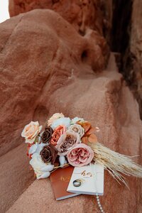 A bouquet of roses in pink, white, and orange hues rests on a red rock surface. Beside it are two wedding rings and a small booklet with cursive text. Captured by Michelle, the scene is set against a rugged, natural rock formation.