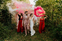 Mariés entourés de demoiselles d'honneur en robe pourpre brandisant des fumigènes rose dans un cadre verdoyant. Photo prise par laura, photographe professionnelle en Vendée.