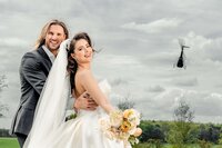 A smiling bride and groom posing outdoors with a bouquet of pastel flowers, as a helicopter flies in the cloudy sky behind them, captured by a UK wedding photographer and videographer.