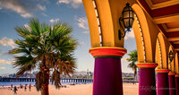 Brightly painted colonnade of the Santa Cruz Beach Boardwalk with beach, palm and wharf in background.