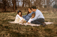 Family of four with mom, dad, and two little girls sit on a blanket in a field with woods behind them. Everyone is looking at one of the little girls while smiling and laughing. 