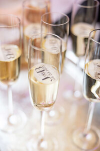 Close-up of champagne flutes filled with bubbly liquid, each glass topped with a circular edible monogram featuring J&B and a date. The glasses are arranged on a reflective surface.