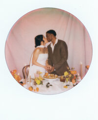Polaroid-style film photo of a bride and groom kissing as they cut their cake at a romantic sweetheart table surrounded by floral decor — a timeless moment captured by a film wedding photographer.