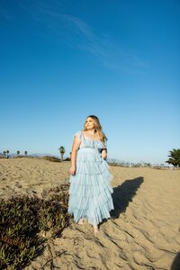 Young woman in a light blue tiered dress standing on a sandy beach under a clear blue sky, with distant palm trees and hazy mountains in the background for a dreamy California vibe.