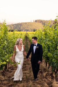 Bride and groom walking through vineyard, holding hands.