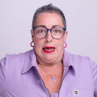 Close-up of Pippa, a person with cropped lilac-grey hair, blue rimmed glasses, and pink hoop earrings wearing lilac collared dungarees, making a wide-eyed, exaggerated “oops” face with bright red lipstick.