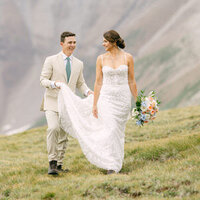 In this touching image, the groom carefully holds up his bride's dress to prevent it from catching on the rocks as they walk through the rugged terrain of the Canadian Rockies. Captured after their private helicopter landing atop a mountain, this moment epitomizes the support and teamwork that define their relationship, set against the breathtaking natural beauty of their wedding backdrop.