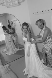 a black and white image of the mother of the bride helping her daughter put on her wedding gown. The bride's dress has a big bow in the back the mother is tying. The bride is wearing silk elbow-length gloves. The photo is film inspired as it's black and white and taken at a dutch angle with direct flash.